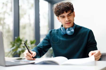 Photo for Concentrated student guy writing down info to his workbook while participating in online webinar or preparing for exam, sitting in coworking space, free space - Royalty Free Image
