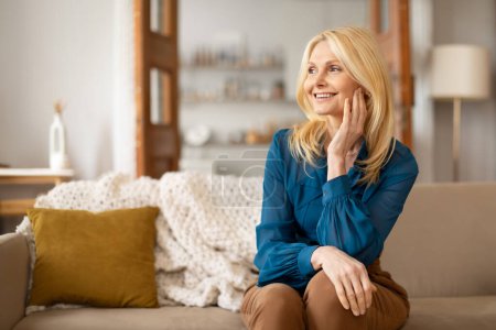 Photo for Attractive Blonde Middle Aged Woman Relaxing On Sofa Indoor, Touching Face With Smooth Skin, Posing In Casual Clothes At Home, Looking Aside And Enjoying Her Weekend In Domestic Comfort - Royalty Free Image