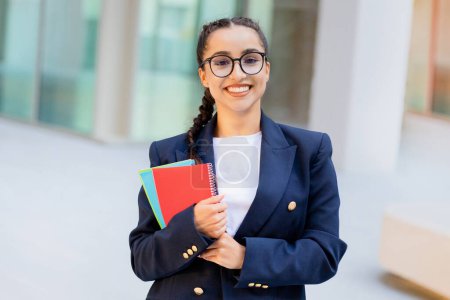 Photo for Cheerful pretty young brunette lady wearing smart casual outfit and eyeglasses student posing at college campus, holding notepads in her hands. Education, school, university concept - Royalty Free Image