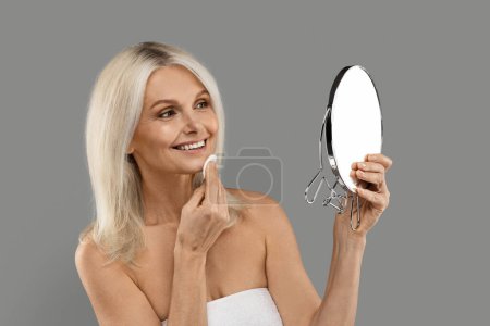 Foto de Hermosa señora mayor mirando en el espejo y la cara de limpieza con almohadilla de algodón, atractiva mujer madura haciendo rutina diaria de cuidado de la piel, disfrutando de tratamientos de belleza, de pie sobre fondo gris - Imagen libre de derechos