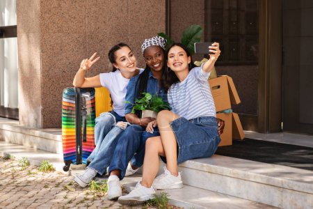 Téléchargez les photos : Jour du déménagement, concept de déménagement. Heureuses jeunes femmes multiraciales copines assises à côté de construire en plein air avec effets personnels valise boîtes en papier, prendre selfie sur smartphone, sourire et gestuelle - en image libre de droit