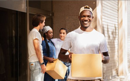 Photo for Happy multicultural group of young friends two guys and two ladies holding boxes with belongings, standing by building, have conversation, student moving to new apartment, relocation - Royalty Free Image