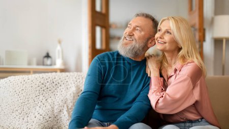 Téléchargez les photos : Souriant romantique conjoints aînés regardant de côté, étreignant et appréciant le confort domestique assis sur le canapé à l'intérieur confortable de la maison moderne. Les conjoints se relaxent ensemble. Panorama, Espace vide - en image libre de droit