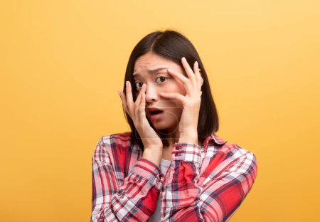 Photo for Scared young chinese lady student in casual outfit covering her face with hands and afraid, standing isolated on yellow background. Human emotions, facial expression and bad news - Royalty Free Image