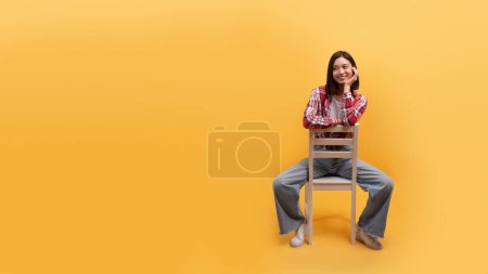 Téléchargez les photos : Jeune Chinoise souriante assise sur une chaise isolée sur fond jaune, studio, panorama avec espace libre. Grande annonce, offre, bonne humeur, repos et détente seul et mode de vie - en image libre de droit