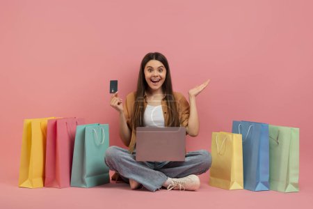 Photo for Big Sales. Excited Teen Girl With Laptop And Credit Card Sitting Among Bright Shopping Bags, Happy Young Woman Enjoying Making Internet Purhcases, Posing Over Pink Studio Background, Copy Space - Royalty Free Image