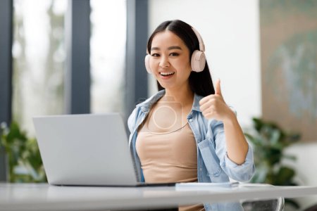 Téléchargez les photos : Heureuse asiatique étudiante recommandant cours d'étude en ligne, montrant pouce levé et souriant à la caméra. Dame dans un casque assis au bureau, faisant comme un geste - en image libre de droit