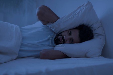 Foto de Problema de sueño. Hombre indio estresado cubriendo la cabeza con almohada mientras está acostado en la cama, joven hombre del este despierto en la noche, molesto por el sonido fuerte, que sufre de insomnio o problemas mentales - Imagen libre de derechos