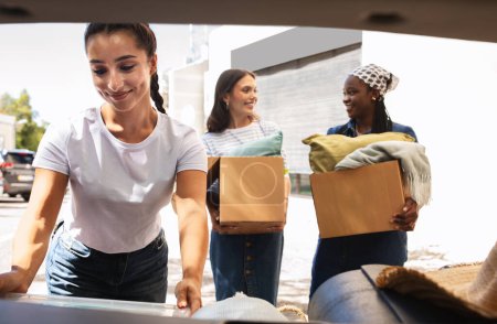 Photo for Moving day, relocation, new house. Three young ladies packing car with cardboxes full of belongings. Happy multiracial women with paper boxes moving to new apartment, leaving dormitory - Royalty Free Image