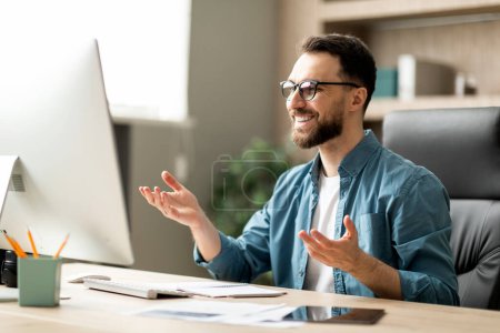 Foto de Hombre atractivo empresario haciendo videollamada mientras está sentado en el escritorio en la oficina, hombre de negocios milenario mirando el monitor de la computadora y sonriendo, disfrutando de la comunicación en línea, primer plano - Imagen libre de derechos
