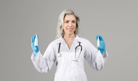 Photo for Woman doctor in white coat showing medical drops bottles in both hands, advertising medicines isolated on gray background in studio, smiling to camera. Gray haired physician recommending treatment - Royalty Free Image
