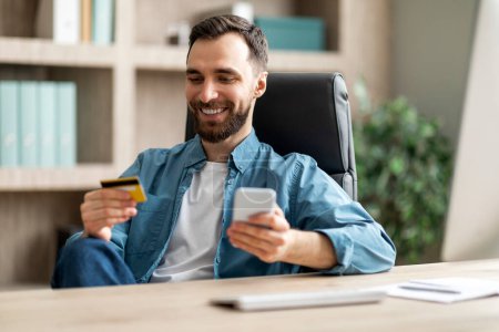 Photo for Online Payment. Smiling Young Businessman Using Smartphone And Credit Card While Sitting At Desk In Office, Handsome Male Entrepreneur Checking Mobile Banking, Browsing Modern App For E-Commerce - Royalty Free Image