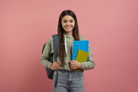 Foto de Retrato de joven estudiante feliz con mochila y libros posando sobre fondo de estudio rosa, hermosa adolescente sosteniendo libros de trabajo y sonriendo a la cámara, disfrutando de la educación moderna, espacio para copiar - Imagen libre de derechos