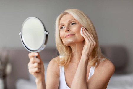 Foto de Hermosa señora rubia de mediana edad mirando en el espejo, borrando maquillaje de su cara, utilizando almohadillas de algodón y loción de limpieza, sentado en el dormitorio, espacio libre - Imagen libre de derechos