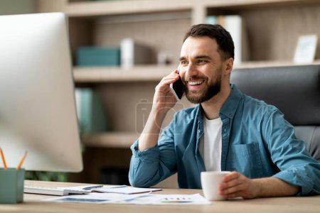 Photo for Mobile Communication. Handsome Male Entrepreneur Talking On Cellphone On Workplace In Office, Millenial Business Man Sitting At Desk With Computer, Enjoying Pleasant Phone Conversation, Closeup Shot - Royalty Free Image