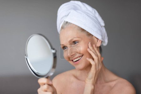 Téléchargez les photos : Concept de traitement du visage. Belle femme d'âge moyen en serviette sur la tête regardant dans le miroir et touchant sa peau douce parfaite, debout dans la salle de bain, espace de copie - en image libre de droit