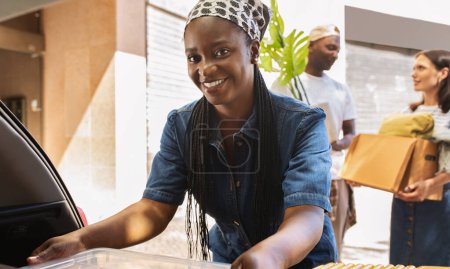 Photo for Cheerful pretty young african american woman packing boxes with her stuff to auto trunk, smiling at camera. Happy multiracial friends helping black lady to move to new apartment - Royalty Free Image