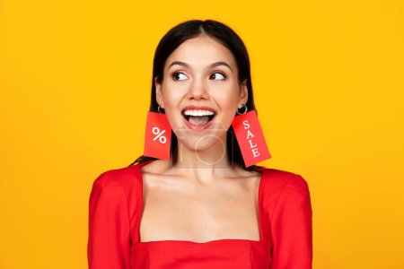 Photo for Black Friday Concept. Portrait Of Happy Excited Woman Wearing Sale Labels As Earrings, Cheerful Young Shopaholic Lady Enjoying Shopping Discounts, Posing Isolated Over Yellow Background, Copy Space - Royalty Free Image