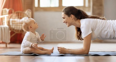 Foto de Vista lateral de la alegre joven madre deportiva haciendo ejercicio con su pequeño bebé en casa. Mamá haciendo ejercicio regular planchado en la alfombra de fitness con lindo hijo bebé rubio, panorama - Imagen libre de derechos
