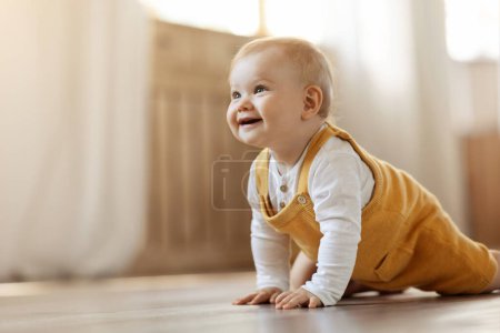 Adorable happy blonde infant baby crawling on floor by home in living room. Portrait of smiling cute child toddler moving on his own by hous, looking at copy space and smiling