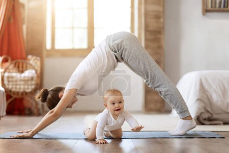 Young mother and her little baby daughter doing yoga exercises on fitness mat at home. Adorable toddler kid crawling next to athletic well-fit mom. Sporty family lifestyle, mutual activities concept