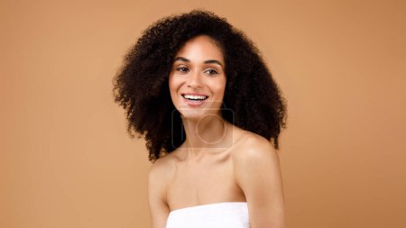 Foto de Belleza natural. Feliz joven latina con el pelo rizado disfrutando de la rutina de autocuidado de pie envuelta en toalla y sonriente, posando sobre fondo marrón del estudio, panorama - Imagen libre de derechos