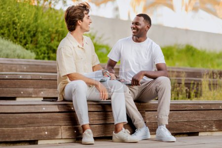 Téléchargez les photos : Deux jeunes hommes étudiants multiraciaux traînant sur le campus universitaire, assis sur le banc à côté de l'université, se préparant pour la classe ensemble, avoir une conversation et sourire, espace de copie - en image libre de droit