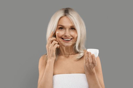 Photo for Antiage Cosmetics. Beautiful Mature Woman Applying Moisturizing Cream On Face, Attractive Senior Female Holding Jar With Nourishing Lotion, Standing Wrapped In Towel Over Grey Background, Copy Space - Royalty Free Image
