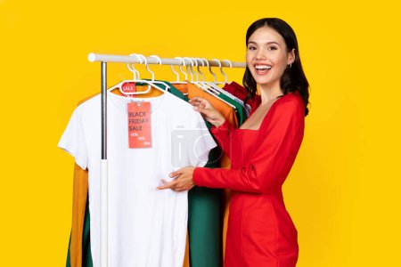 Photo for Happy Excited Young Woman Pointing At Clothes With Red Sale Tag, Cheerful Beautiful Lady Enjoying Seasonal Sales And Discounts, Standing Near Clothing Rack Isolated Over Yellow Background - Royalty Free Image