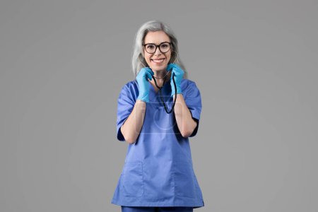 Téléchargez les photos : Dame infirmière souriante posant avec stéthoscope portant uniforme bleu et lunettes debout sur fond gris. Portrait d'un travailleur médical confiant prêt pour un rendez-vous médical et un bilan de santé - en image libre de droit