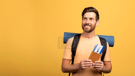 Téléchargez les photos : Joyeux homme d'âge moyen avec un sac à dos contenant un passeport international et des billets d'avion, regardant de côté l'espace libre sur fond de studio jaune. Voyageur masculin prêt à partir en vacances à l'étranger - en image libre de droit