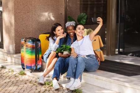 Photo for Cheerful attractive young multiethnic ladies helping their girlfriend moving to new house, sitting on stairs outdoor next to building with boxes, taking selfie on cell phone while waiting for taxi - Royalty Free Image