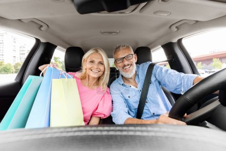 Photo for Cheerful beautiful elderly spouses driving home after shopping in mall, smiling. Happy senior man and woman sitting inside car, holding colorful shopping bags purchases. Black friday, season sale - Royalty Free Image