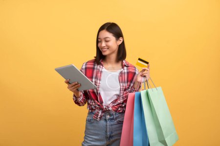 Téléchargez les photos : Joyeux jeune femme asiatique avec des sacs-cadeaux à l'aide d'un ordinateur tablette et carte de crédit pour acheter des choses en ligne, faire du shopping dans la boutique en ligne sur fond de studio jaune - en image libre de droit