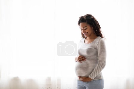 Foto de Concepto de embarazo. Retrato de una joven mujer embarazada negra sonriente abrazando el vientre mientras estaba de pie cerca de la ventana en casa, una mujer afro-americana embarazada tocando tiernamente su barriga, Copiar el espacio - Imagen libre de derechos