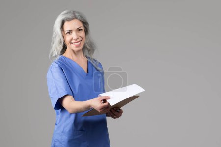 Téléchargez les photos : Femme infirmière souriante en tenue de travail bleue avec stéthoscope et dossier médical posant sur fond de studio gris. Bannière Web avec médecin femme tenant presse-papiers, espace vide pour l'annonce de texte - en image libre de droit