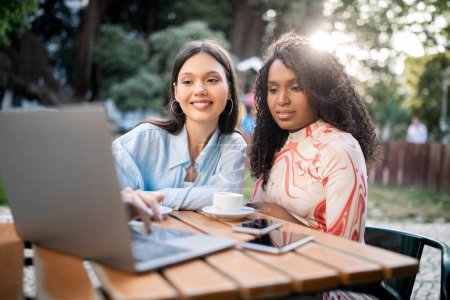 Photo for Two multiethnic female friends using laptop and drinking coffee at cafe terrace, smiling beautiful young women shopping online or browsing new website on computer while relaxing outdoors, closeup - Royalty Free Image