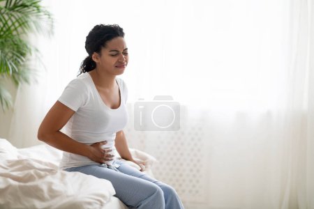 Foto de Ache de estómago. Mujer negra enferma que sufre de dolor abdominal agudo en el hogar, afligida mujer afroamericana que tiene dolor menstrual o problemas de digestión, sentada en la cama y tocando el vientre, espacio copiado - Imagen libre de derechos