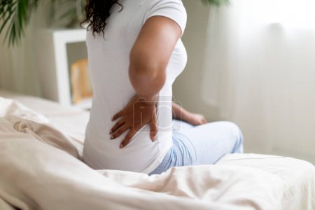 Téléchargez les photos : Jeune femme noire ayant le dos contre terre tout en étant assise sur le lit à la maison, Zone de douleur de massage féminine afro-américaine méconnaissable, Toucher le bas du dos, Inflammation du rein, Prise de vue coupée - en image libre de droit