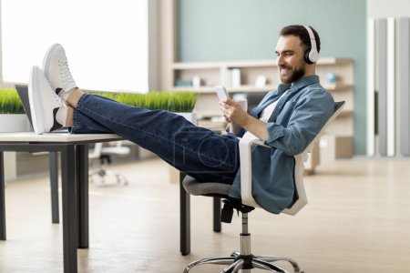 Téléchargez les photos : Profiter du temps libre. Homme souriant se relaxant au travail au bureau, reposant les jambes sur la table et utilisant un smartphone, employé masculin heureux penché sur la chaise et utilisant le téléphone mobile, prenant une pause du travail - en image libre de droit