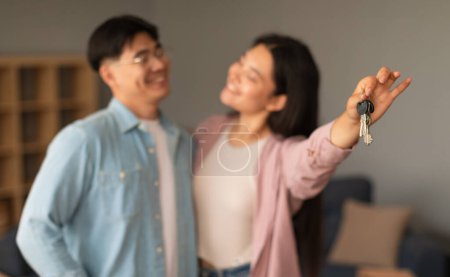 Téléchargez les photos : Propriétaires immobiliers. Joyeux couple japonais tenant de nouvelles clés de maison, les montrant à la caméra debout à l'intérieur. Jeunes conjoints Publicité Propriété Location ou vente Offre. Focus sélectif sur la clé - en image libre de droit