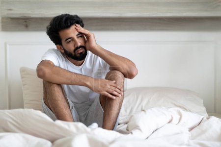 Photo for Fatigue Concept. Tired Young Indian Man Sitting In Bed At Home, Exhausted Upset Millennial Eastern Guy Waking Up In Bedroom, Feeling Unwell, Having Headache Or Suffering Insomnia, Free Space - Royalty Free Image