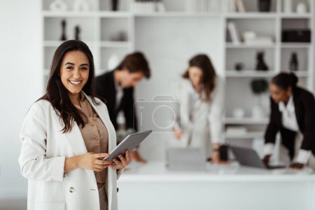 Photo for Latin businesswoman ceo posing with digital tablet in office interior, smiling at camera during business meeting with her colleagues. Career success - Royalty Free Image