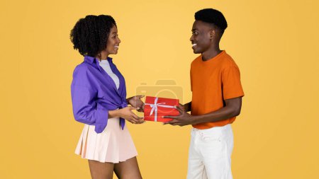 Photo for Cheerful millennial black lady gives gift box to man, congratulate, isolated on yellow studio background. Birthday greeting, anniversary surprise, holiday celebration together - Royalty Free Image
