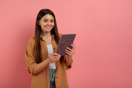 Téléchargez les photos : Concept d'éducation en ligne. Adolescente souriante tenant une tablette numérique et souriant à la caméra, adolescente heureuse utilisant un gadget moderne pour l'étude à distance, posant sur fond de studio rose, espace de copie - en image libre de droit