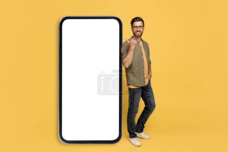 Photo for Cheerful middle aged man leaning and pointing at big smartphone with white blank screen, smiling at camera, showing free space for advertisement, standing over yellow background, mockup - Royalty Free Image