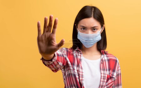 Photo for Confident japanese female student in protective mask showing palm, stop sign with hand, isolated on yellow background, studio shot. Health problems, ill, viruses concept - Royalty Free Image