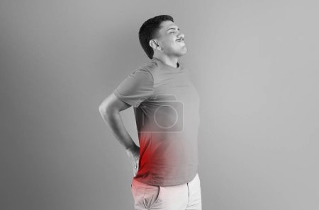 Téléchargez les photos : Homme brésilien touchant le bas du dos avec une tache rouge sur la colonne vertébrale, souffrant de douleur, debout sur le fond du studio, photo en noir et blanc - en image libre de droit