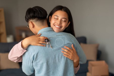Photo for Real Estate Dream Comes True. Happy Asian Couple Hugging, Wife Holding New House Keys, Celebrating Property Purchase At Home Interior, Standing Among Cardboard Moving Boxes - Royalty Free Image