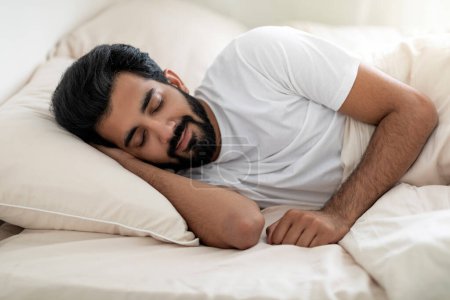 Foto de Retrato de un joven guapo indio que duerme en una cama cómoda en casa, primer plano de la calma Millennial hombre oriental durmiendo en un acogedor dormitorio, acostado sobre una almohada suave con la mano debajo de la cabeza - Imagen libre de derechos